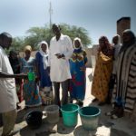Un homme en blouse blanche apprend à des femmes en habits traditionnels à traiter l'eau pour la rendre potable devant un centre de santé au Tchad
